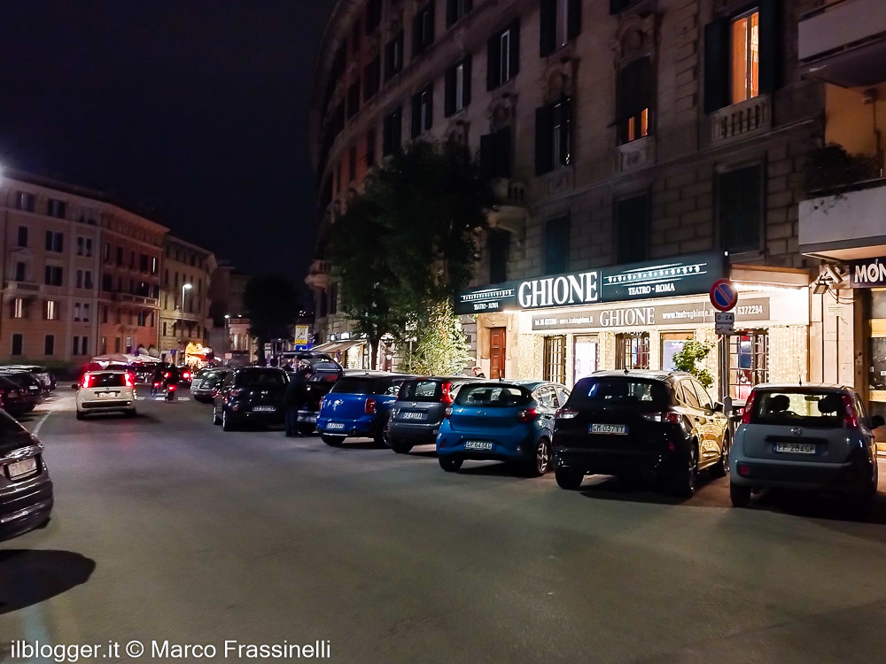 Veduta notturna di una strada di Roma con l'insegna luminosa del Teatro Ghione che brilla tra le auto parcheggiate e i palazzi storici
